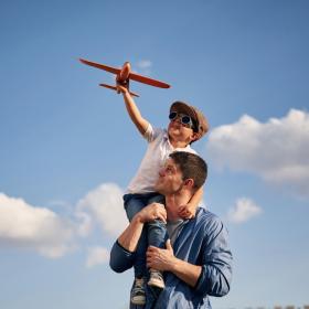 אבא מרים את הבן על הכתפיים. הבן מחזיק מטוס ומשקפי טייס